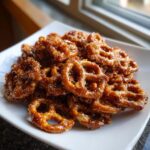 A close-up of a pile of shiny, glazed toffee pretzels coated in a rich caramel mixture, served on a white plate.