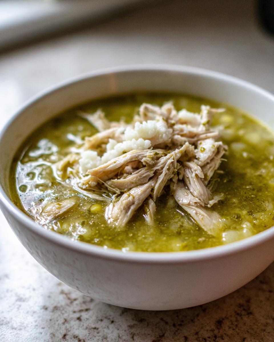 Close-up of a white bowl filled with green chicken pozole, topped with shredded chicken and cotija cheese.