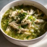 Close-up of a white bowl filled with vibrant green chicken pozole, topped with shredded chicken and fresh cilantro.