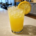 A tall glass filled with bright yellow, iced lemonade, garnished with a fresh lemon slice on the rim.