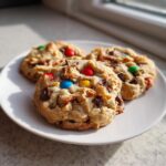 Three freshly baked kitchen sink cookies loaded with chocolate chips and colorful M&Ms sitting on a white plate.