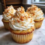 Three moist coconut cupcakes topped with white frosting and toasted coconut flakes.