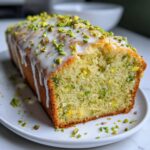 A close-up of a freshly baked pistachio bread loaf, drizzled with white glaze and topped with chopped pistachios.