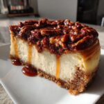 A decadent slice of pecan pie cheesecake topped with glazed pecans and caramel dripping down the side.