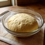 A large, risen ball of light yellow pizza dough recipe resting in a clear glass bowl.