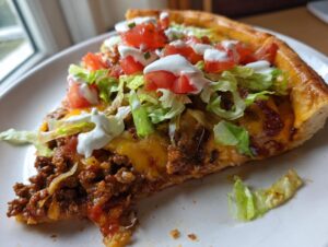 A close-up slice of taco pizza loaded with seasoned beef, melted cheese, lettuce, tomatoes, and sour cream.