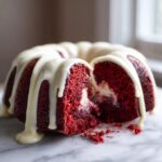 A slice cut from a vibrant red velvet bundt cake drizzled with white glaze, revealing a cream cheese filling.
