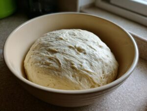A large, risen ball of pizza dough recipe resting in a beige ceramic bowl, ready for shaping.