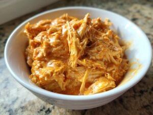 Close-up of shredded chicken coated in a creamy orange buffalo sauce, ready for slow cooker buffalo chicken chili.