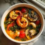 Close-up overhead shot of a bowl of vibrant Tom Yum soup featuring large shrimp, mushrooms, and cherry tomatoes.