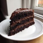 A decadent slice of three-layer chocolate fudge cake covered in thick, glossy chocolate frosting.