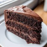 A close-up of a rich, dark slice of homemade chocolate cake layered with chocolate frosting.