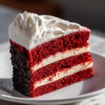 A decadent slice of red velvet cheesecake showing three layers of red cake separated by white cream cheese filling and topped with whipped cream.