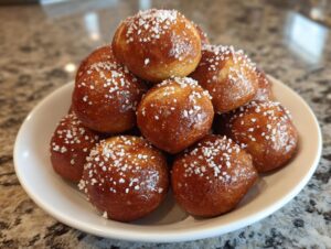 A mound of freshly baked, golden brown soft pretzel bites topped with coarse white salt, served on a white plate.