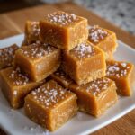 A stack of soft, square salted caramels sprinkled with flaky sea salt on a white plate.