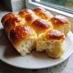 A plate of golden brown sourdough discard rolls, one split open showing the fluffy interior and shiny glaze.