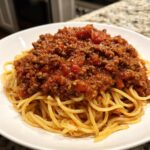 A close-up of spaghetti noodles generously topped with a thick, rich bolognese sauce in a white bowl.