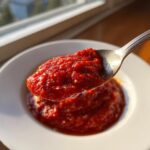 A spoonful of thick, bright red, no-cook pizza sauce being lifted from a white bowl.