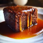 Close-up of a rich, dark square slice of sticky toffee pudding drenched in warm toffee sauce.