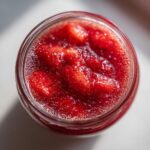 Overhead close-up view of bright red strawberry freezer jam with visible fruit chunks inside a glass jar.