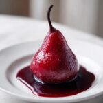 A single, glistening, deep red poached pear served on a white plate with a pool of rich poaching liquid.