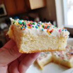A hand holding up a thick, moist slice of sugar cookie bars topped with white frosting and colorful sprinkles.