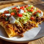 A close-up of a slice of taco pizza topped with seasoned ground beef, melted cheese, lettuce, tomatoes, and sour cream.