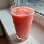 A tall glass filled with a vibrant, icy watermelon smoothie sitting on a sunlit windowsill.