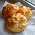 Three freshly baked, golden brown cloud bread rounds resting on white parchment paper near a window.