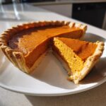 A close-up of a whole vegan pumpkin pie with one slice cut out and resting beside it on a white plate.