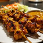 Close-up of perfectly grilled, golden brown chicken satay skewers served with peanut sauce and a side of pickled vegetables.