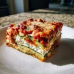A thick, layered slice of zucchini lasagna featuring zucchini noodles, white ricotta cheese, and red marinara sauce, topped with melted mozzarella.