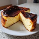 A creamy, burnt-top Basque cheesecake, with one slice cut and resting next to the main cake on a white plate.