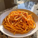 A bowl of bucatini pasta coated in vibrant orange butternut squash pasta sauce, topped with grated cheese and black pepper.