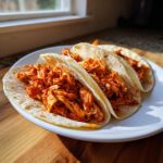 Three soft corn tortillas filled generously with vibrant, saucy chicken tinga, served on a white plate.