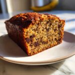 A close-up of a moist slice of vegan banana bread with visible chocolate chips, served on a white plate.