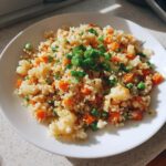 A white plate filled with bright cauliflower rice mixed with diced carrots, peas, and topped with fresh green onions.