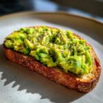 Close-up of creamy avocado toast topped with black pepper and seeds, highlighting the texture.