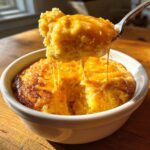 A spoonful of creamy, bright yellow cheese grits being lifted from a white bowl, showing long, gooey cheese pulls.