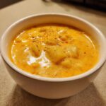 Close-up of a bowl of rich, orange creamy cheese soup filled with visible chunks of potato and topped with black pepper.