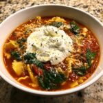 Close-up of a hearty bowl of creamy lasagna soup featuring pasta, spinach, tomato broth, and a large dollop of ricotta cheese.