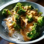 Close-up of rice noodles topped with bright green broccoli florets smothered in creamy peanut sauce.