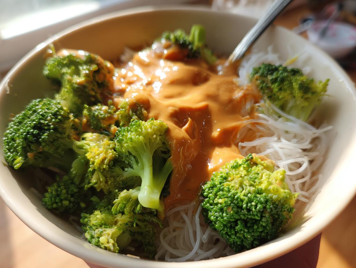 A bowl of rice noodles topped with bright green broccoli florets and generously covered in creamy peanut sauce.