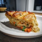 A close-up of a creamy slice of turkey pot pie recipe showing flaky golden crust, carrots, and peas.