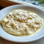 A close-up of thick, creamy white pizza sauce seasoned generously with black pepper and dried herbs in a white bowl.