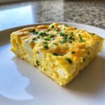 A square slice of fluffy sheet pan eggs, featuring visible white cheese curds and topped with fresh green chives.