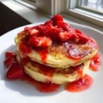 Close-up of a stack of fluffy strawberry pancakes generously topped with macerated strawberries and syrup.