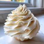 Close-up of a perfectly piped swirl of fluffy vanilla buttercream frosting on top of a small cake or cupcake base.