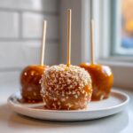 Three homemade caramel apples on a plate, one coated in sparkling sugar crystals.