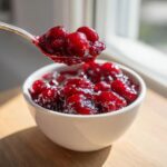 A spoonful of glossy, thick cherry pie filling being lifted from a white bowl.